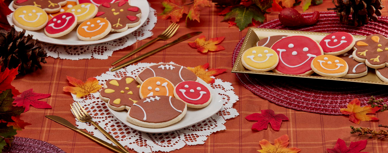 Festive Thanksgiving cookies, including turkey cookies, football cookies, and smiley face cookies, displayed on autumn-themed plates and tablecloth with fall leaves and pinecones.