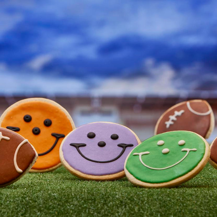 an array of smiley face cookies decorating in football team colors along with football shaped cookies on a football field backdrop