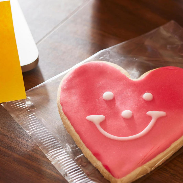 A heart shaped sugar cookie decorated with a cheerful smile amidst a backdrop of a busy desk with a post it note saying Just Thinking About You