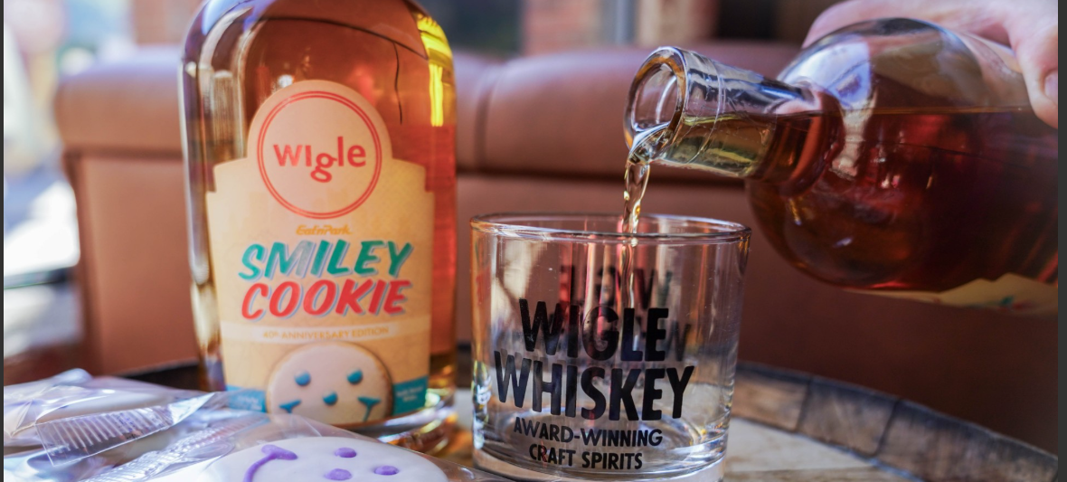 A bottle of Smiley Cookie Wigle Whiskey pouring into a glass on a table full of smiley cookies