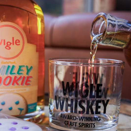 A bottle of Smiley Cookie Wigle Whiskey pouring into a glass on a table full of smiley cookies