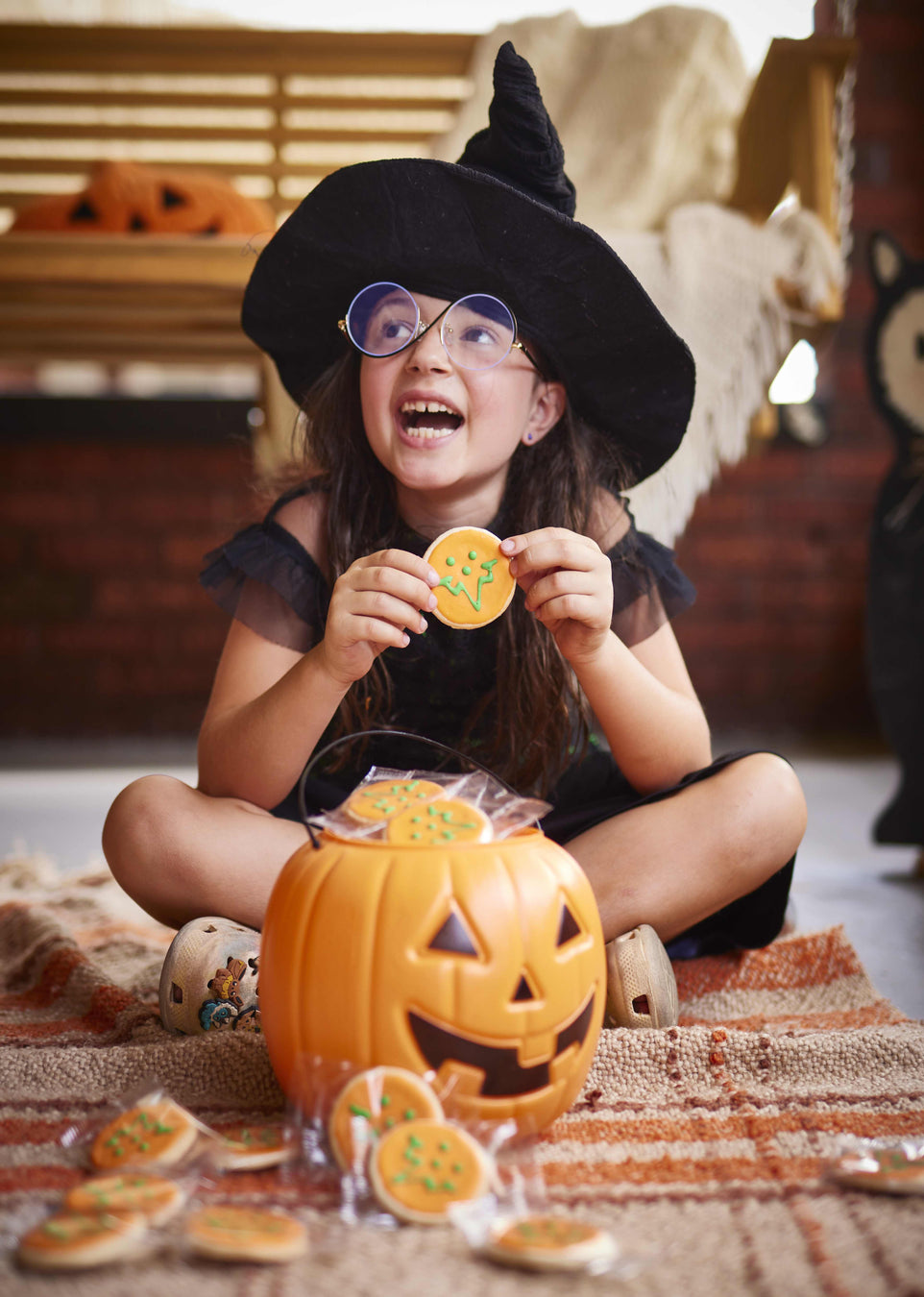Child in Halloween costume with witch hat and glasses, holding a cookie near a pumpkin-shaped candy container.